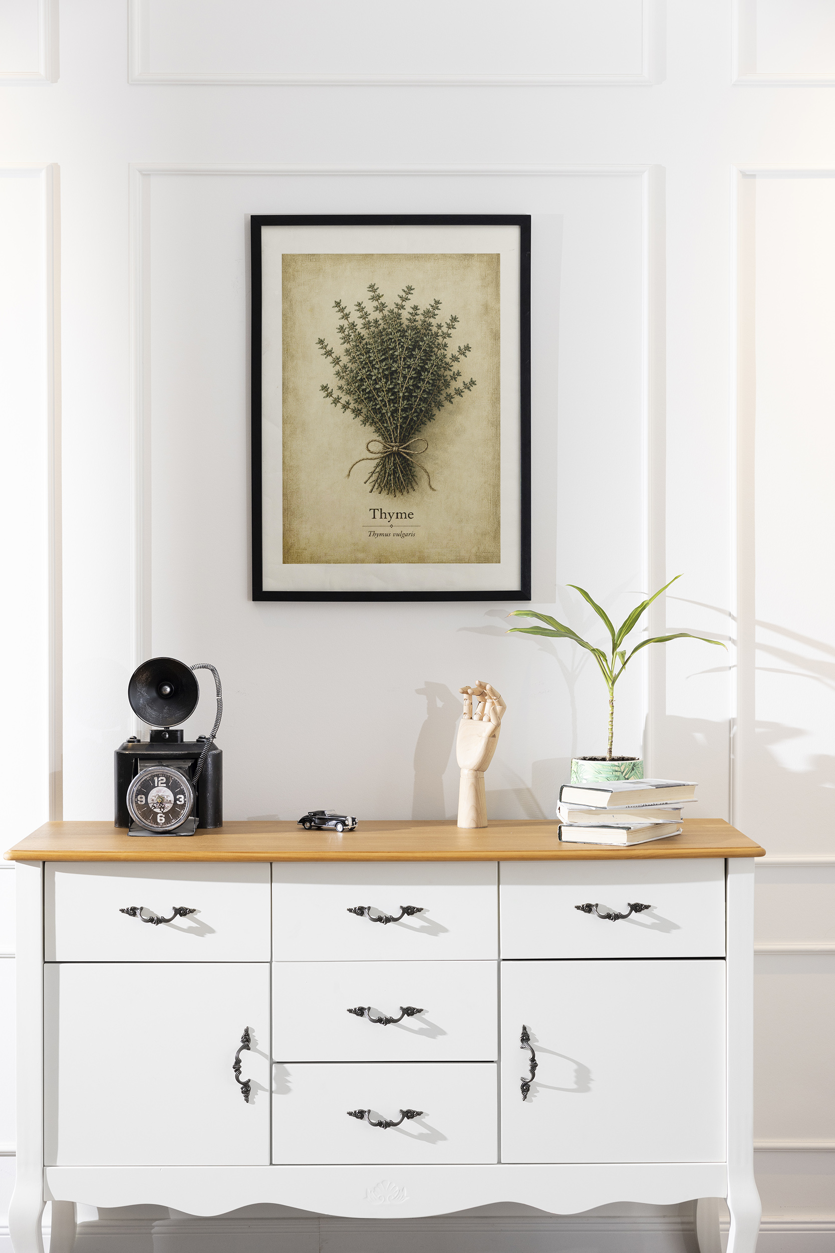 A framed thyme poster with a tied herb bundle and beige parchment background hangs on a white paneled wall above a white console table with books and a small plant.
