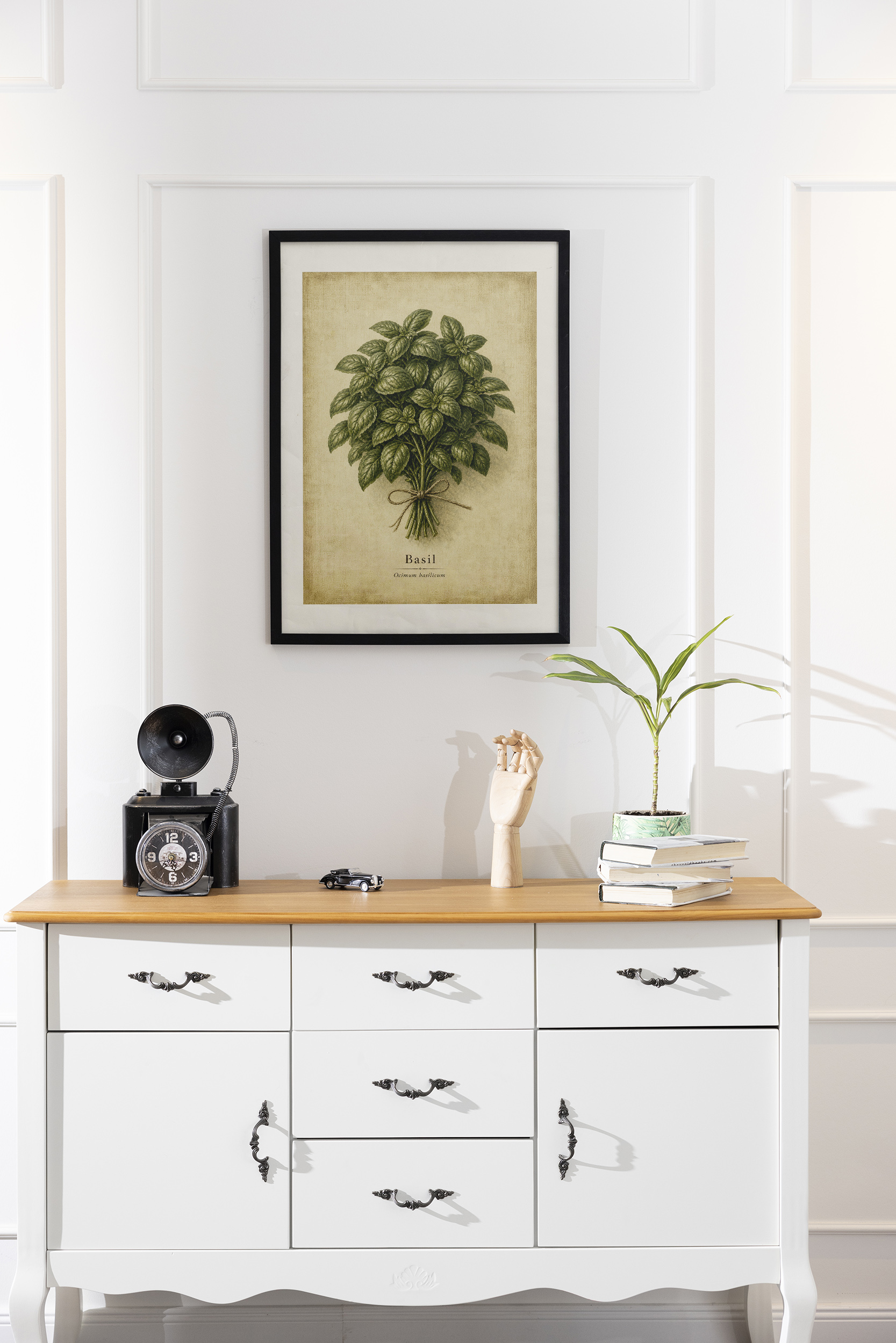 A framed basil poster with a full herb bundle and beige parchment background hangs on a white paneled wall above a white console table with books and a small plant.