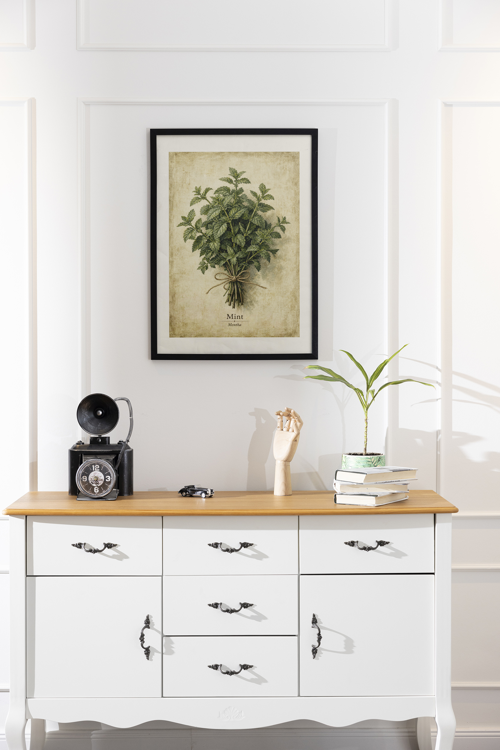 A framed mint poster with a leafy herb bundle and beige parchment background hangs on a white paneled wall above a white console table with books and a small plant.